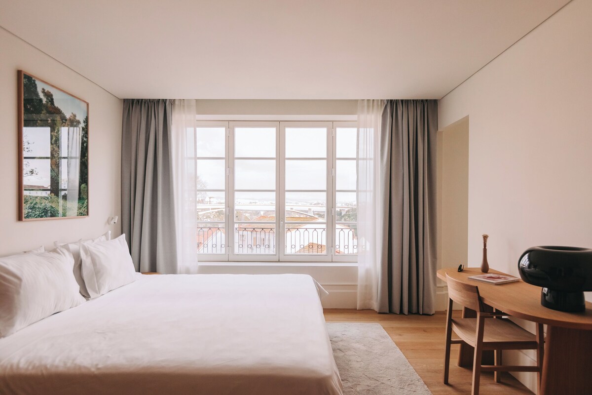 A hotel room with a large bed, a desk with a chair, and a view of buildings through a window.