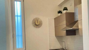 Een compacte keuken met een ingebouwde kookplaat, een spoelbak en witte kastjes. Er is een raam met zonwering, een decoratieve wandklok en een kleine tafel met een tafelkleed met bloemen.