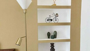 Een kamer met een hoge vloerlamp, een bruine bank en een wandplank met diverse decoratieve objecten.
