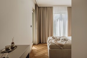 A modern hotel room with a neatly made bed, a bedside table with a candle, and a view of the outdoors through sheer curtains.