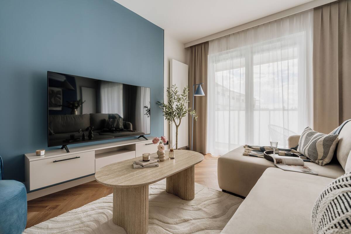 A modern living room with a flat-screen TV, a coffee table, a sofa, and a large window with sheer curtains.