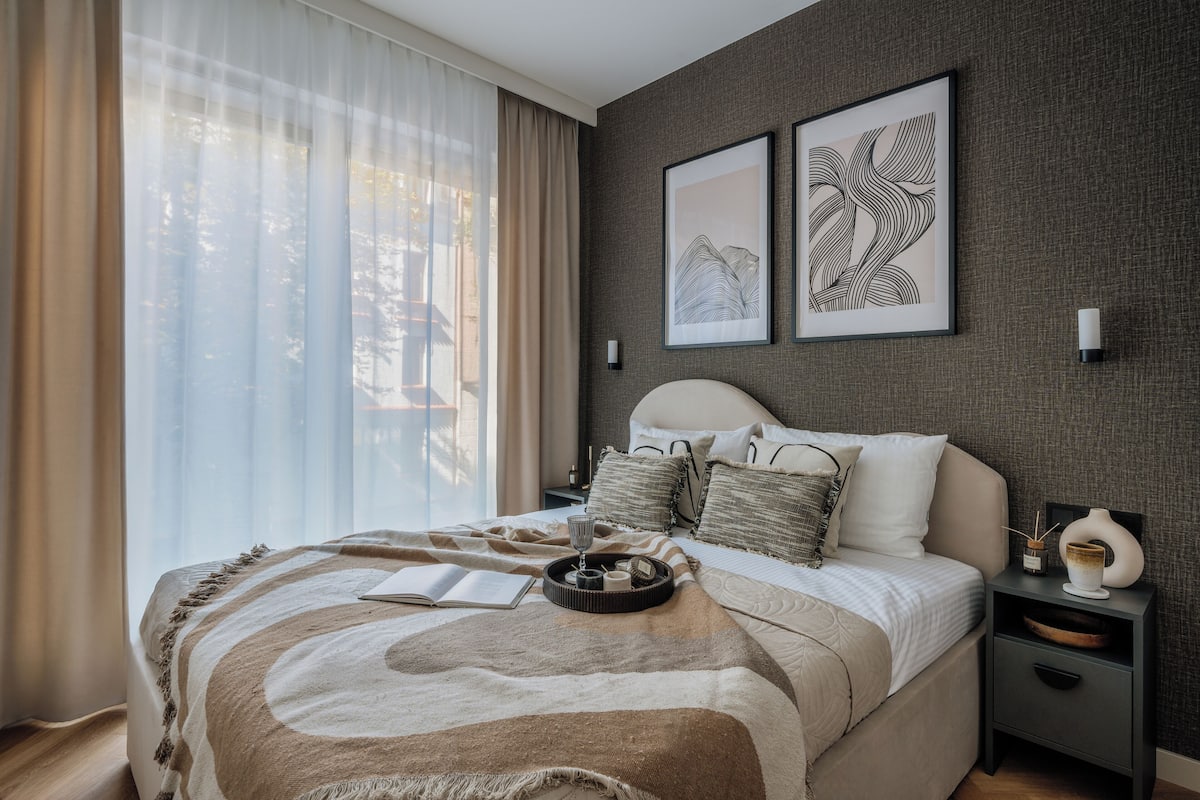 A bedroom with a bed, a bedside table, a nightstand, and a window with sheer curtains.