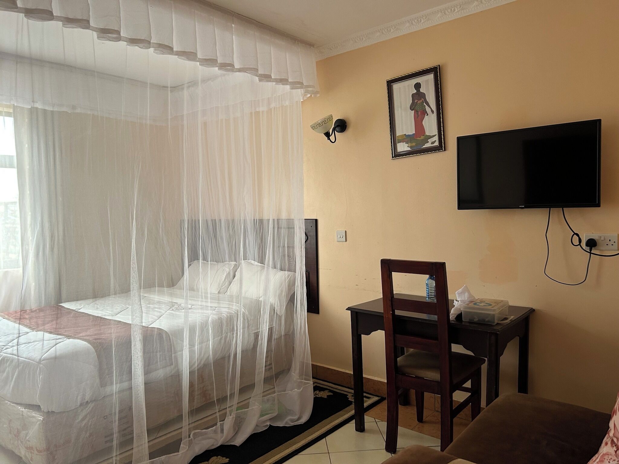 A bedroom with a bed covered by a mosquito net, a desk with a chair, a television on the wall, and a framed picture.