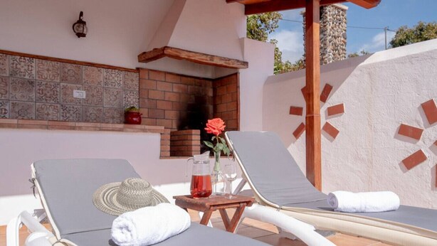 Un área de descanso al aire libre iluminada por el sol, con dos sillones reclinables, una mesita con una jofaina y vasos, un sombrero tejido y una rosa.