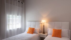 Habitación de hotel con dos camas, una mesita de noche con lámpara y cortinas translúcidas.