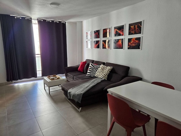A modern living room with a sofa, a small table, and a dining table. There are curtains on the window and artwork on the wall.
