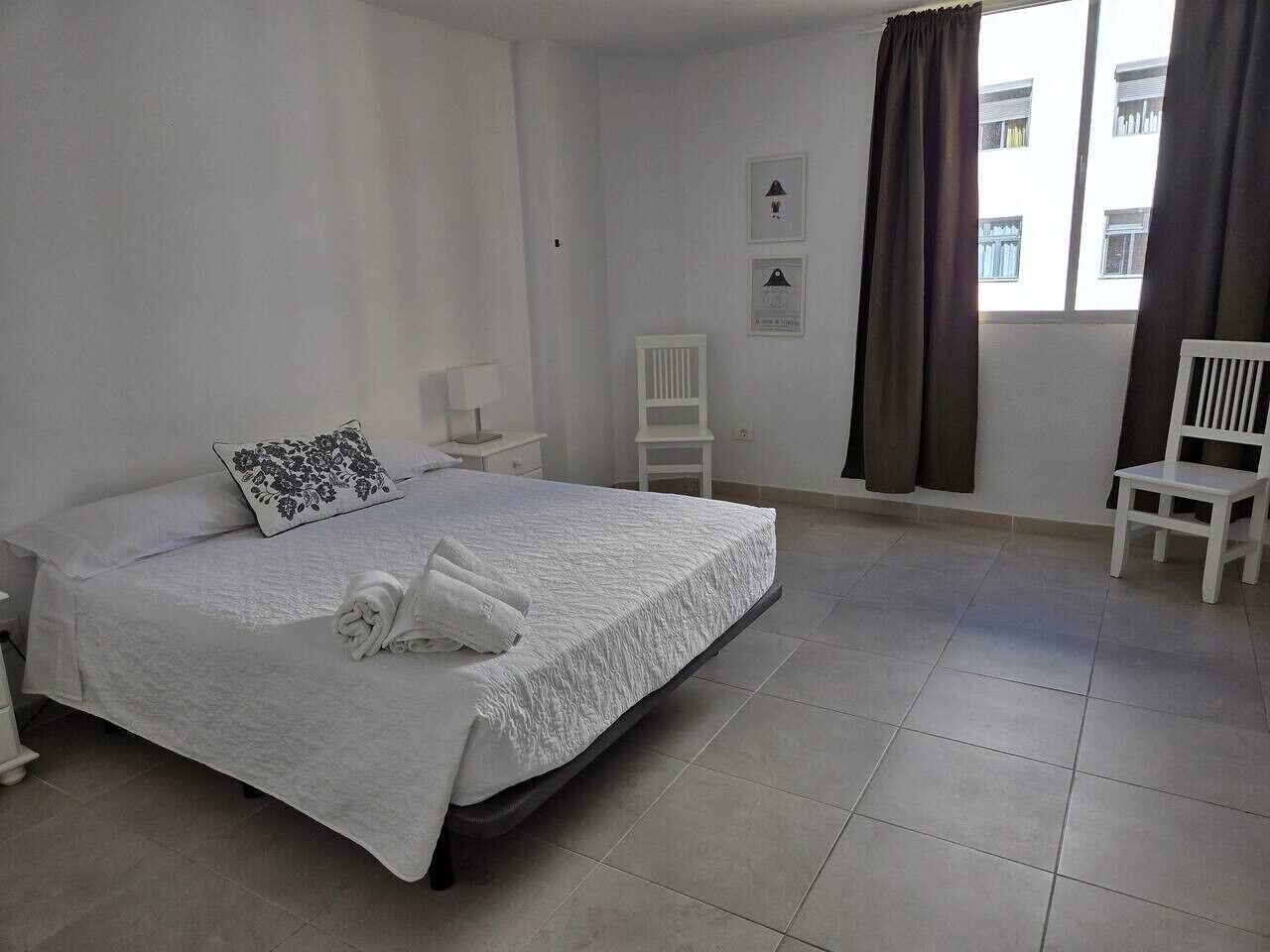 A neatly made bed with a white bedspread and a decorative pillow, a white chair, a small table with a lamp, and a window with curtains.