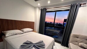 Ett modernt hotellrum med en stor säng, utsikt över stadsbilden i skymningen, en sänggavel i trä och en beige fåtölj.