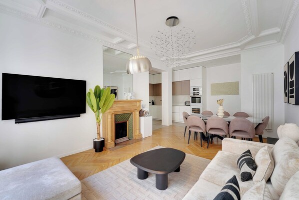 A modern living room with a flat-screen TV, a fireplace, a dining area with pink chairs, and a large mirror.
