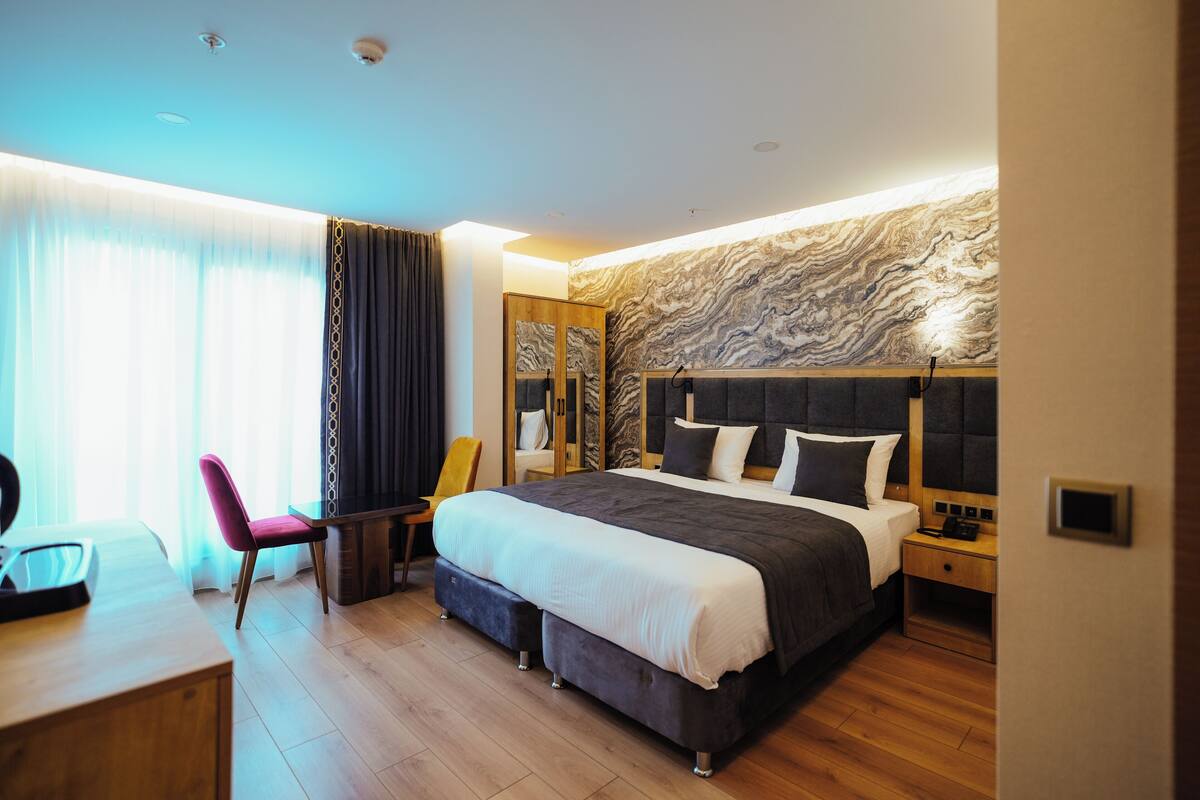 A modern hotel room with a large bed, a desk, a chair, and a wardrobe.