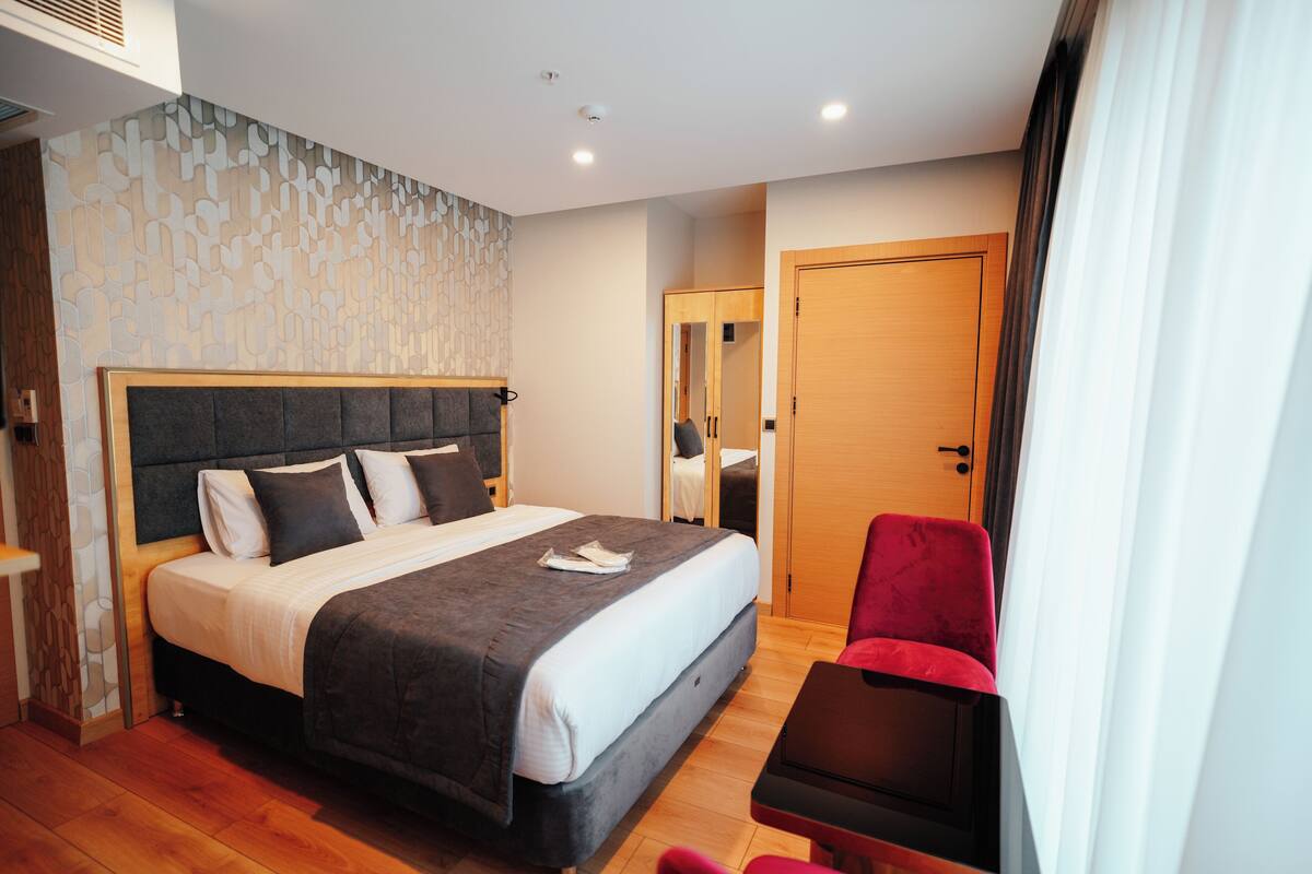 A modern hotel room with a large bed, a red chair, and a wooden wardrobe.