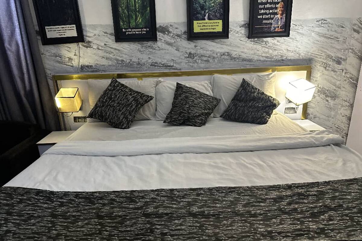 Un lit bien fait, avec du linge de lit blanc et trois coussins à motifs foncés. Deux lampes murales sont fixées de part et d’autre du lit. Au-dessus du lit, trois citations encadrées sont accrochées au mur.