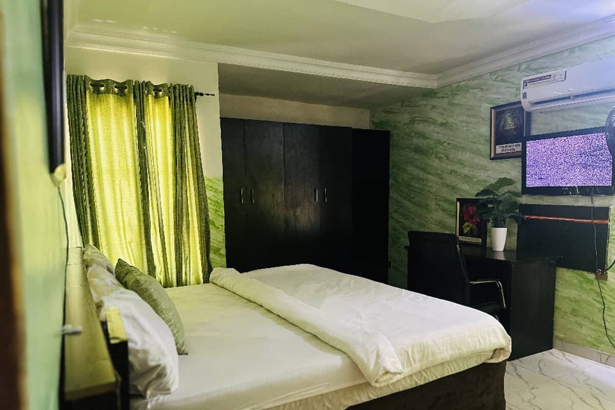Une chambre à coucher avec un lit, une armoire sombre, un bureau avec un ordinateur et un téléviseur fixé au mur.