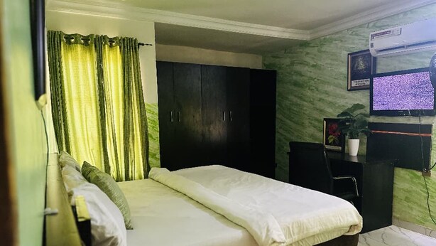 A bedroom with a bed, a dark wardrobe, a desk with a computer, and a television on the wall. - The Autograph Basics Hotel (Lagos)