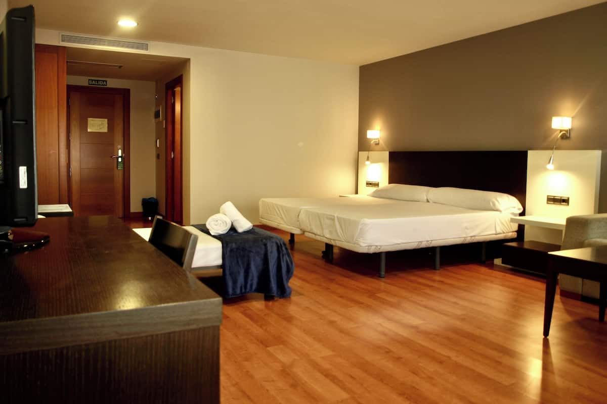 Habitación de hotel con piso de madera, un escritorio con silla, una cama con dos almohadas, una mesita de noche y una lámpara de pared.