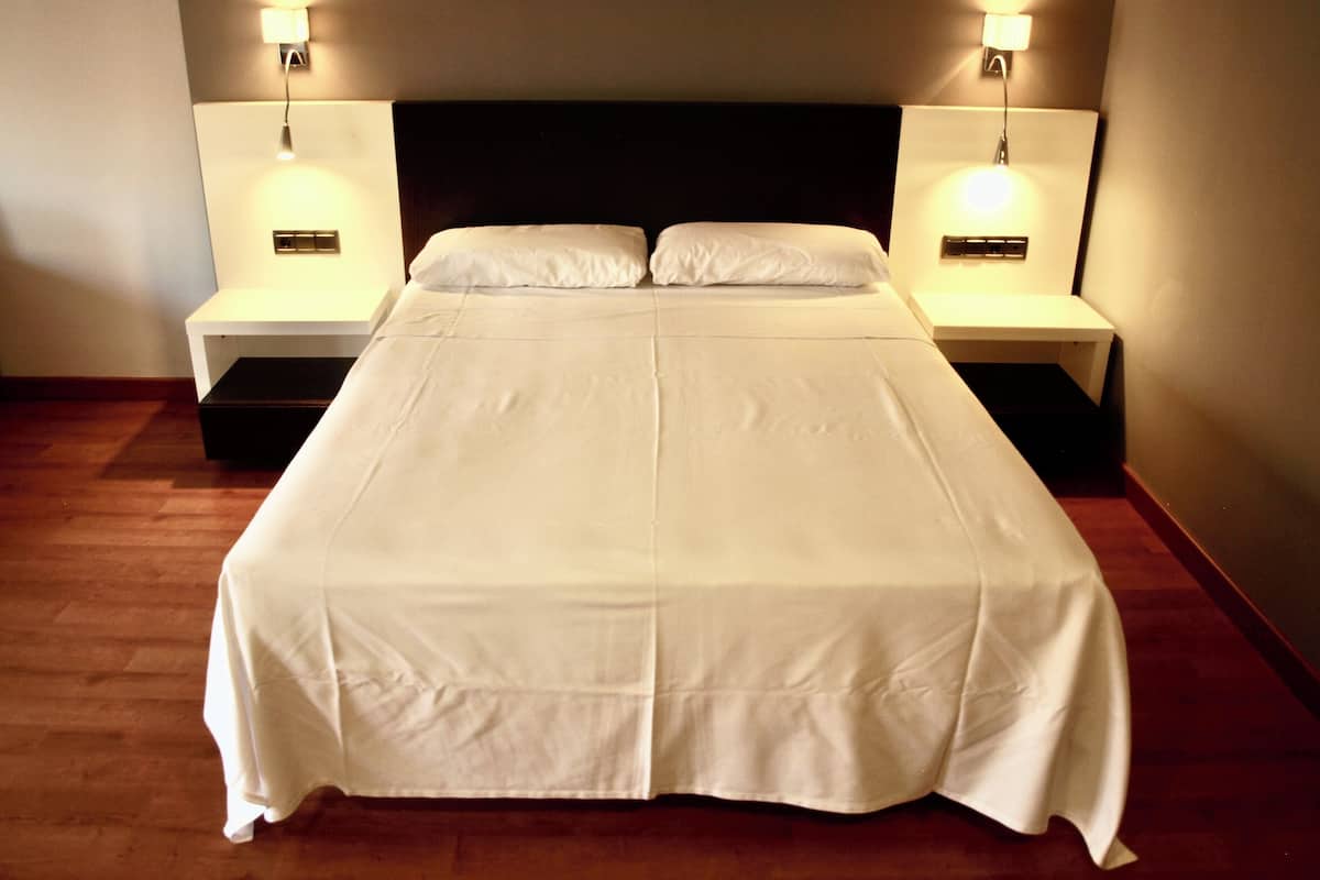 Una cama doble con una colcha blanca, dos mesitas de noche con lámparas y un cabecero oscuro.