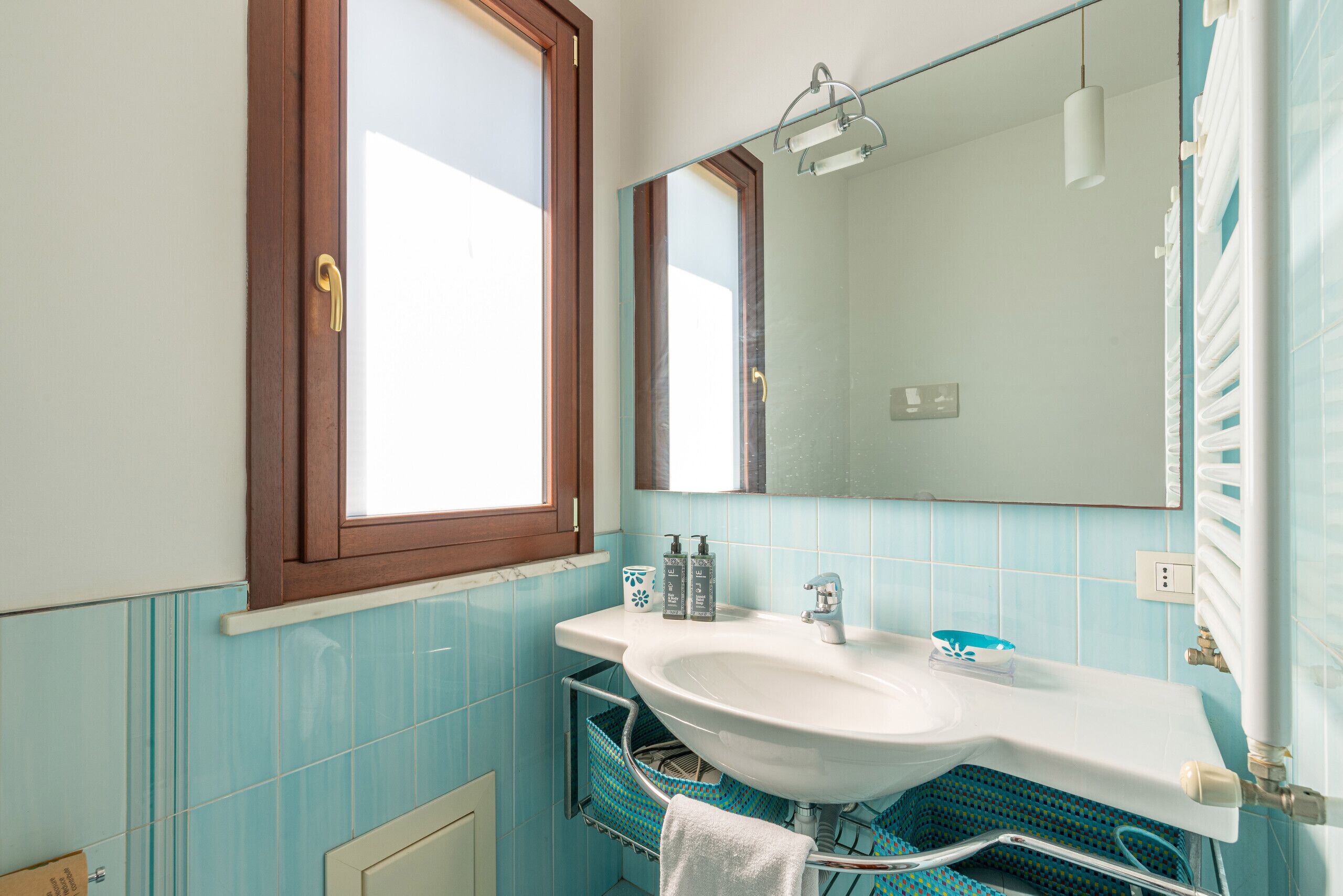 A bathroom with a white sink, blue tiles, a window with a wooden frame, and a mirror above the sink.