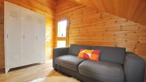 Une pièce aux murs en bois, avec un canapé gris agrémenté d’un coussin coloré, une armoire blanche et un tapis en fourrure sur le sol.
