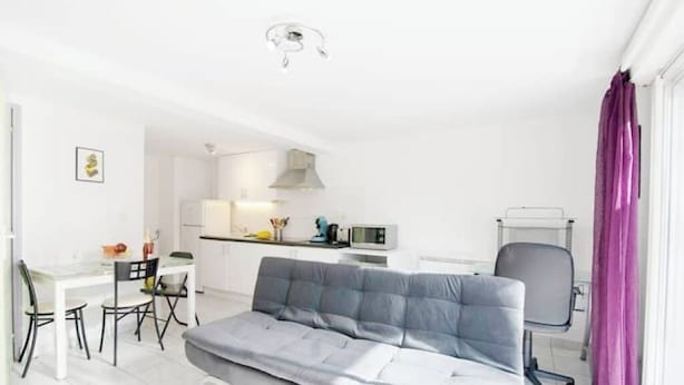 A compact living space with a sofa, dining table, chairs, a kitchenette, and a window with purple curtains.