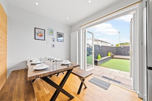 Un comedor con una mesa de madera dispuesta para cuatro, un banco y vistas a un patio trasero con césped y una valla de madera.