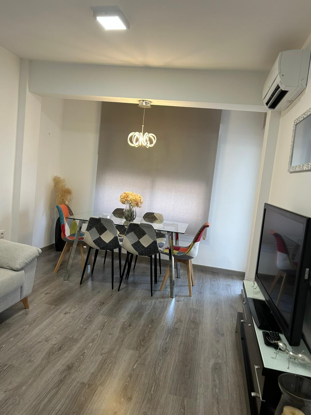 Un salon moderne avec un coin repas, comprenant une table en verre, des chaises à motifs, un lustre, un téléviseur à écran plat et un appareil de climatisation.