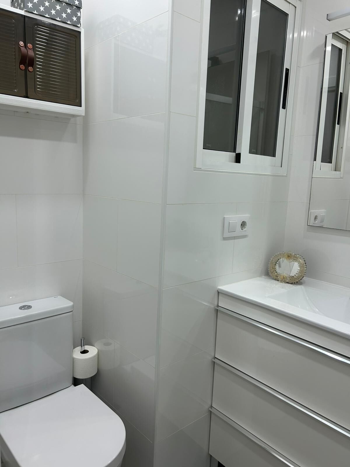 Une salle de bain avec des carreaux blancs, des toilettes, un lavabo avec un miroir et un porte-rouleau de papier toilette.