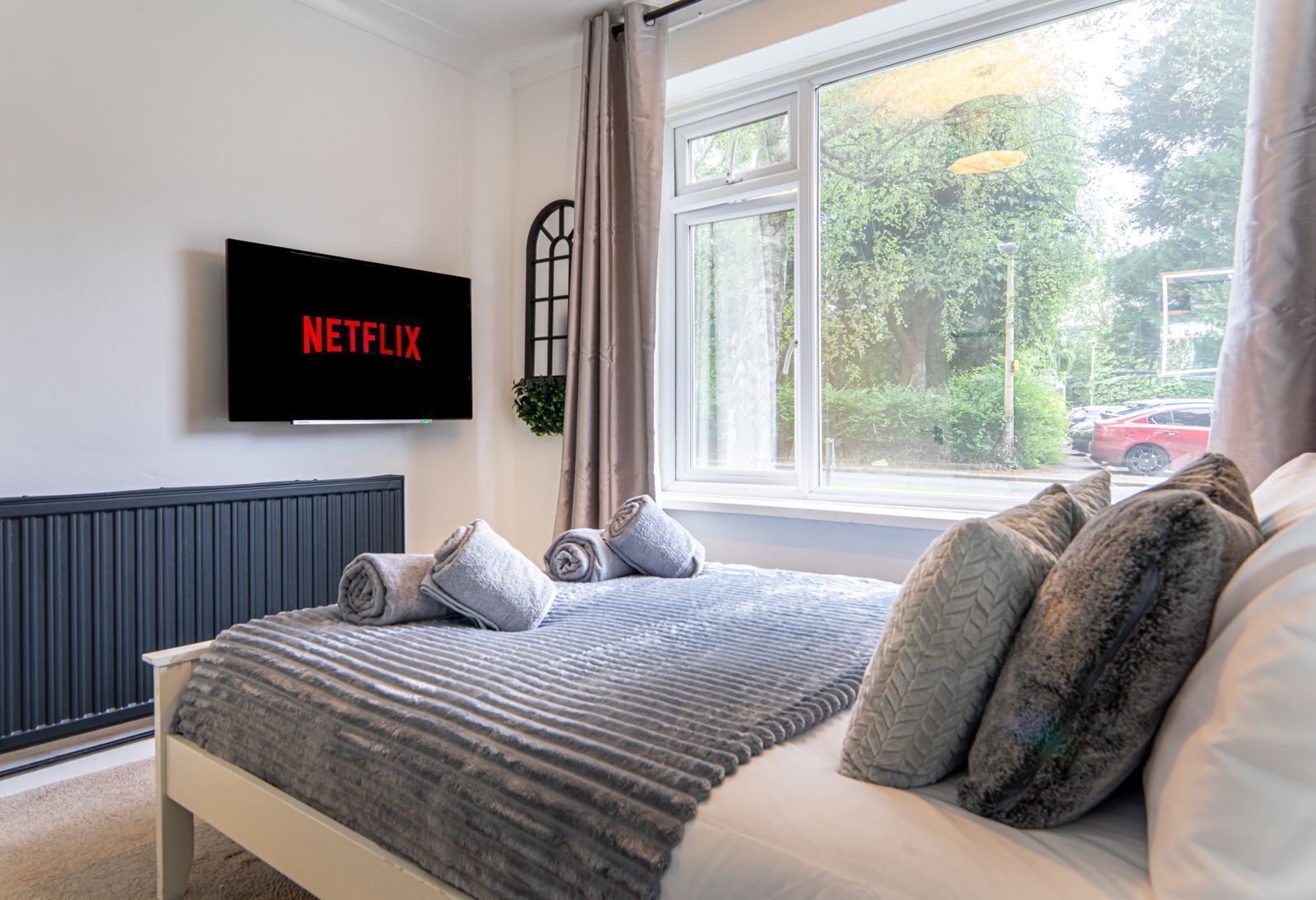 Un dormitori amb un llit, almoinos, un televisor que mostra 'NETFLIX', una finestra amb vistes a vegetació i un cotxe vermell a l'exterior, i un radiador sota el televisor.