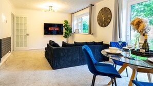 Ein modernes Wohnzimmer mit einem dunkelblauen Sofa, einem Glastisch mit blauen Stühlen, einer Wanduhr, einem Flachbildfernseher mit der Anzeige "NETFLIX" und einem großen Fenster mit Blick auf Grünflächen.