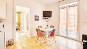 Una habitación luminosa con una mesa blanca y sillas de colores, un televisor en la pared y una puerta corrediza de vidrio al exterior.