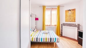 Una habitación pequeña y luminosa con una cama, un radiador, una mesita y un cuadro en la pared.