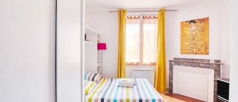 Une petite pièce lumineuse, avec un lit, un radiateur, une petite table et un tableau accroché au mur.