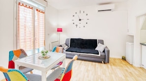 Ein modernes Wohnzimmer mit einem Glastisch, bunten Stühlen, einem grauen Sofa und einer schwarzen Decke.