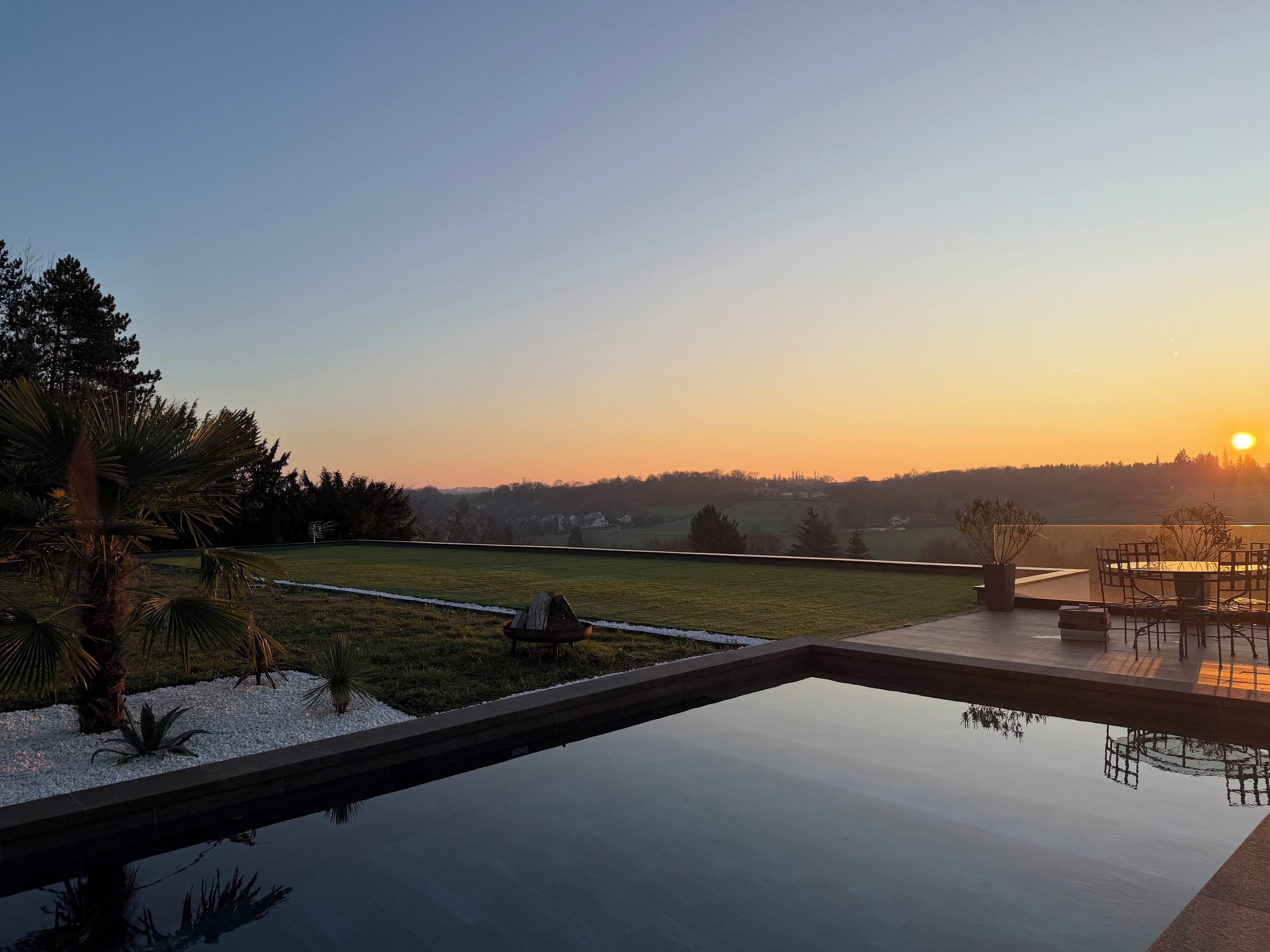 Eine friedliche Außenanlage mit Schwimmbad, einer Terrasse und Blick auf eine ländliche Landschaft bei Sonnenuntergang.