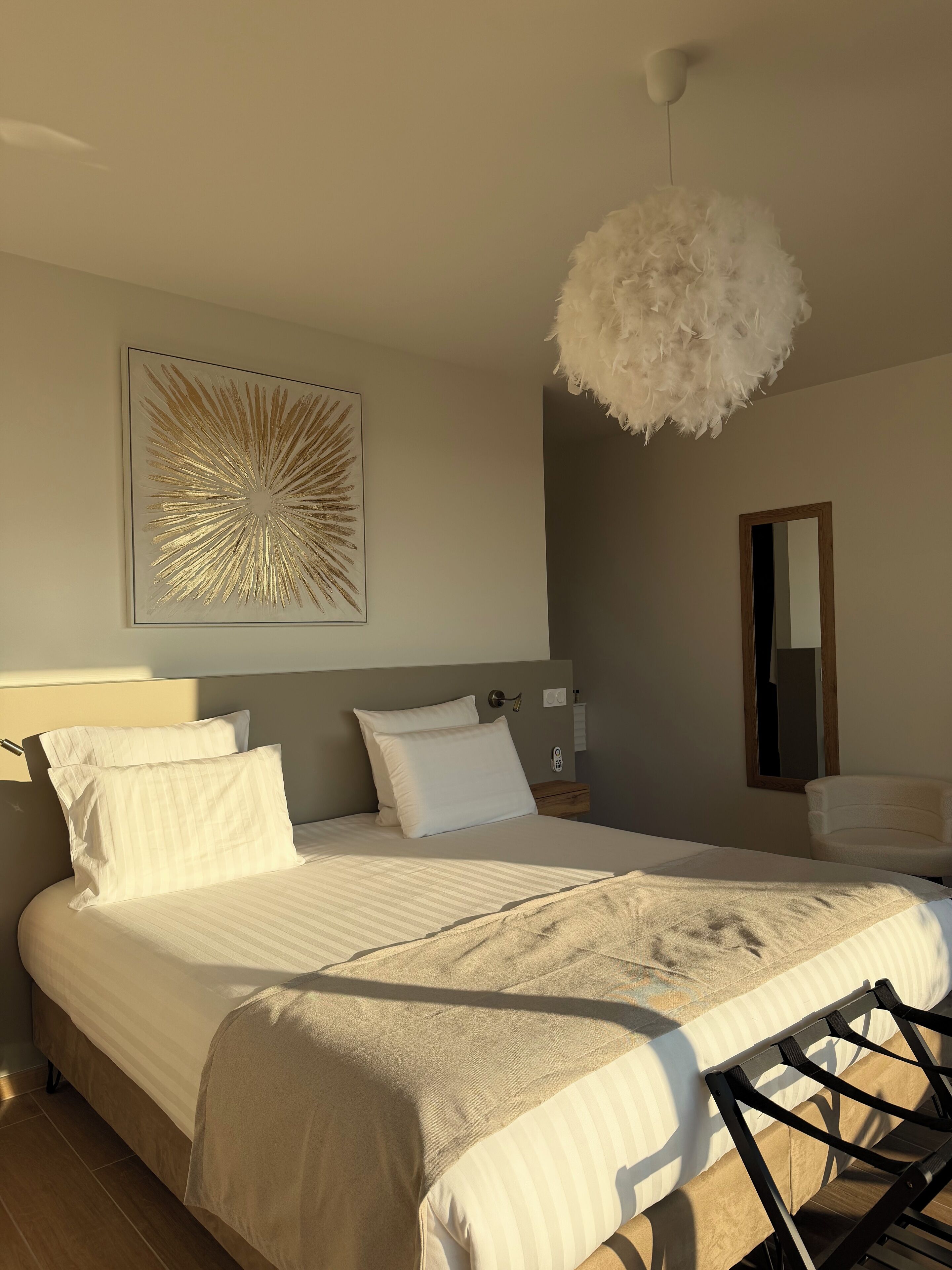 A modern bedroom with a large bed, a feathered light fixture, a framed mirror, and a wall art piece.