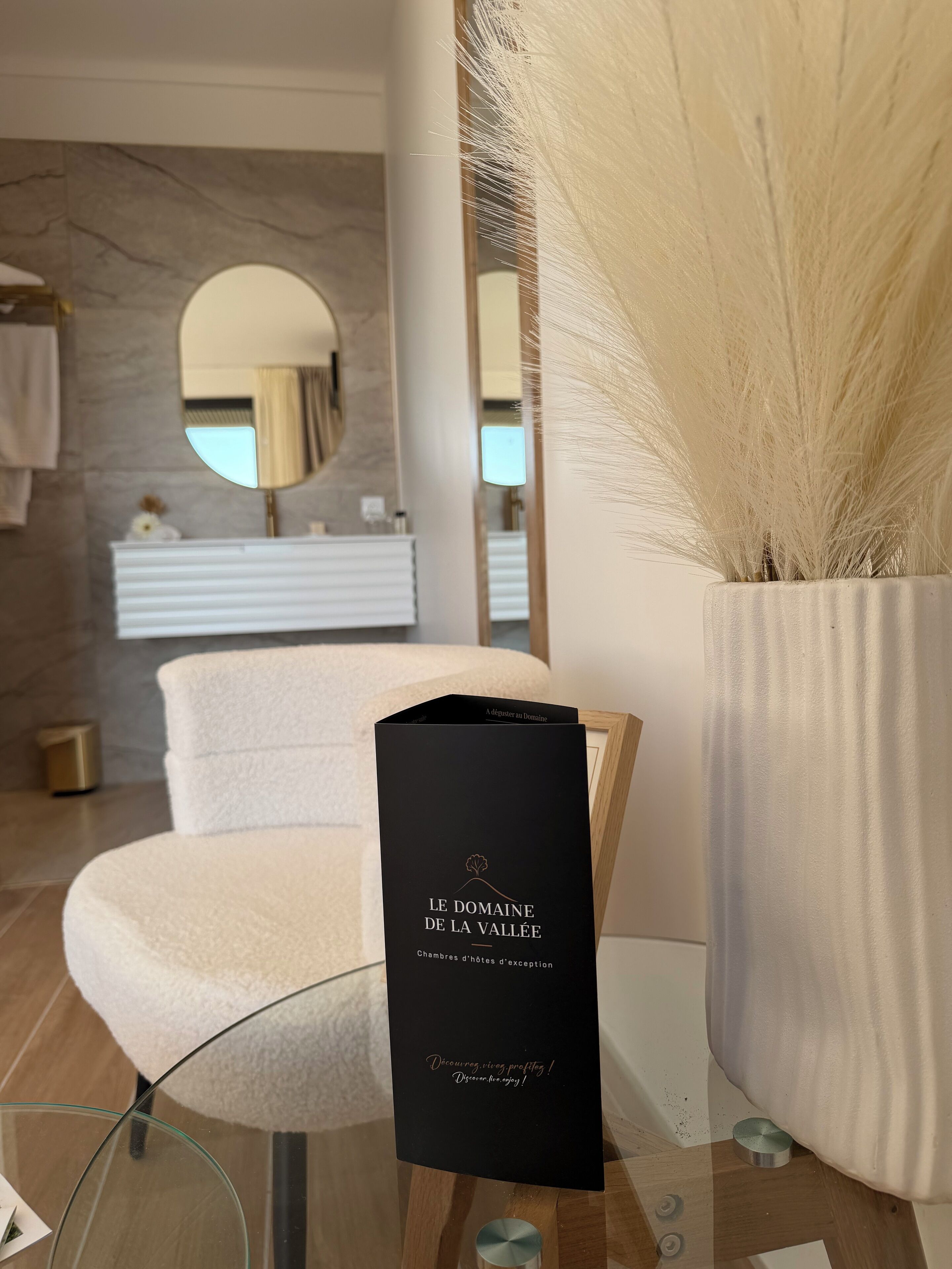Un baño moderno con pared de mármol, un espejo redondo, una toalla blanca y una bolsa de regalo negra sobre una mesa de vidrio.