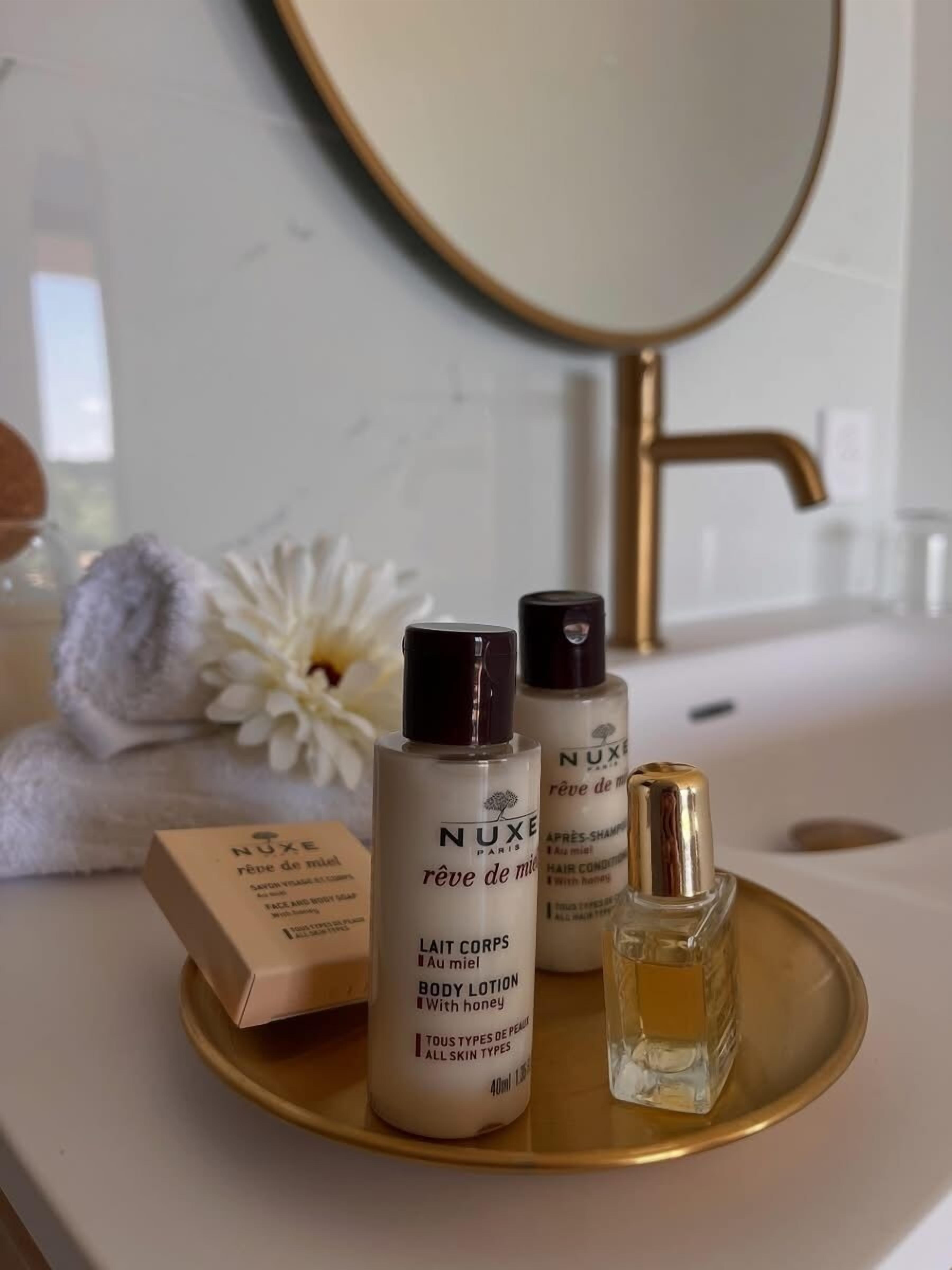 A bathroom vanity with a white sink, two Nuxe body lotion bottles, a small perfume bottle, and a white flower.