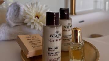 A bathroom vanity with a white sink, two Nuxe body lotion bottles, a small perfume bottle, and a white flower.