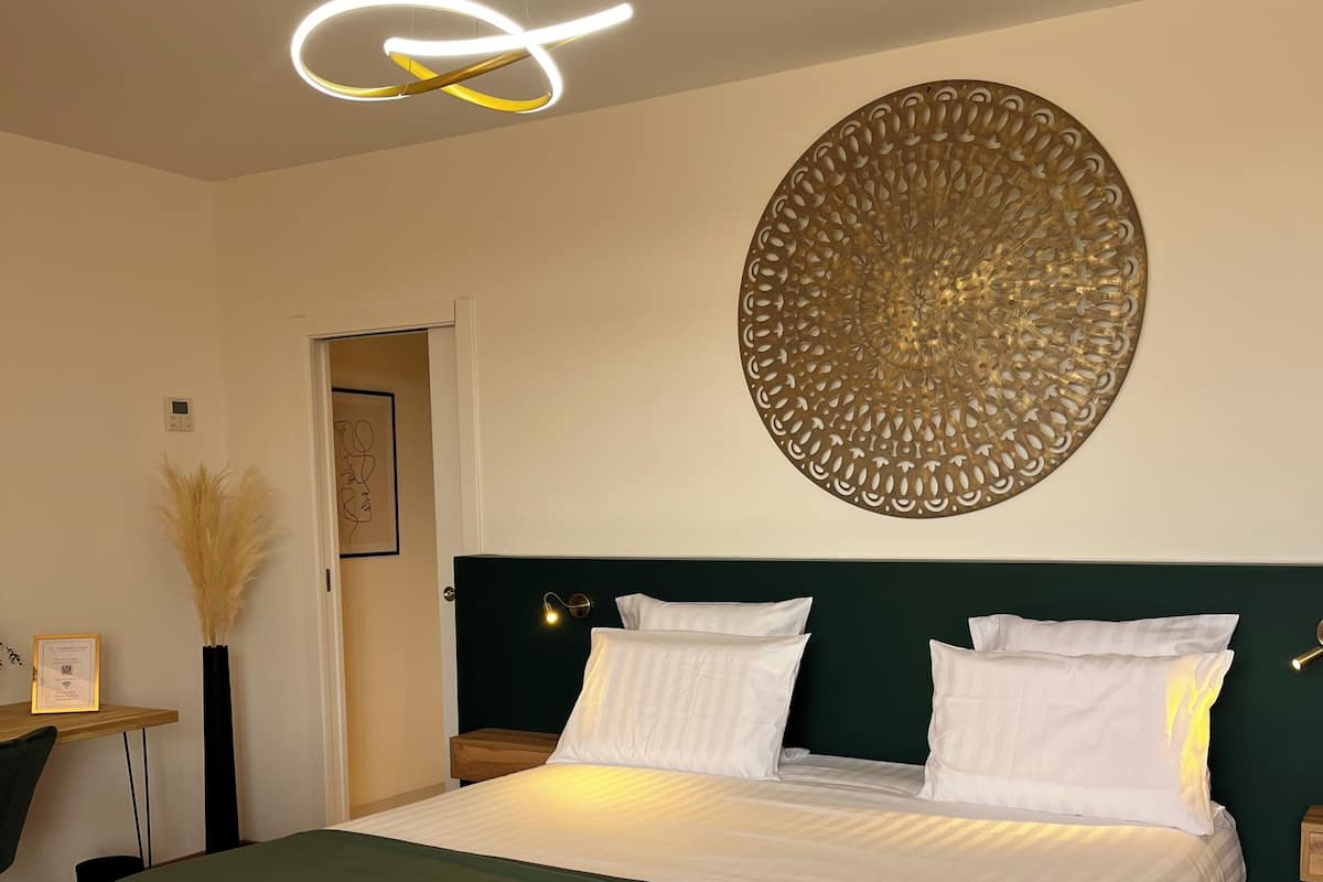 Un lit bien fait, avec une tête de lit rembourrée de couleur verte, une décoration murale et un luminaire moderne au-dessus.