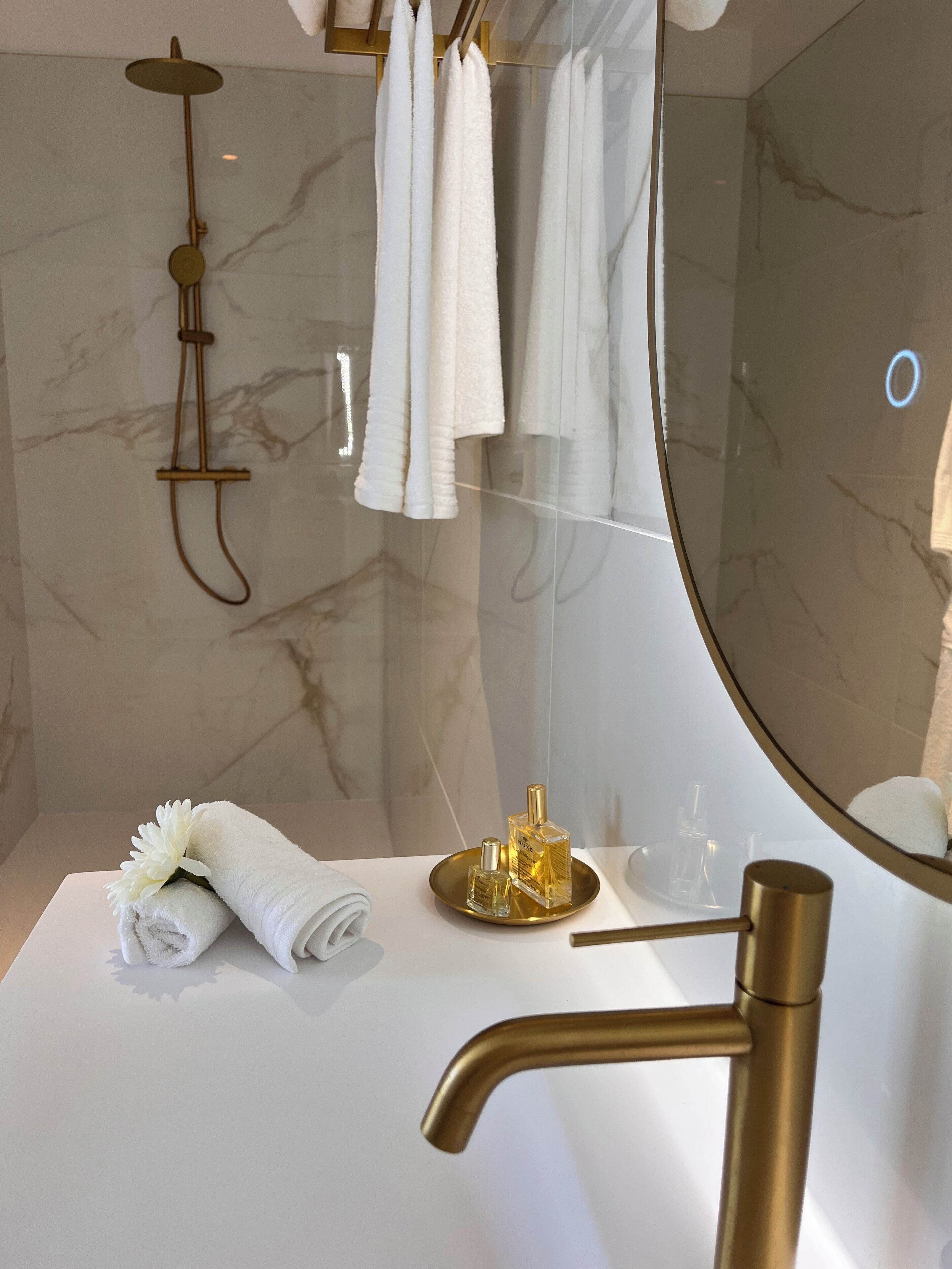 A modern bathroom with a marble wall, a gold showerhead, a large mirror, and a white sink with a gold faucet.