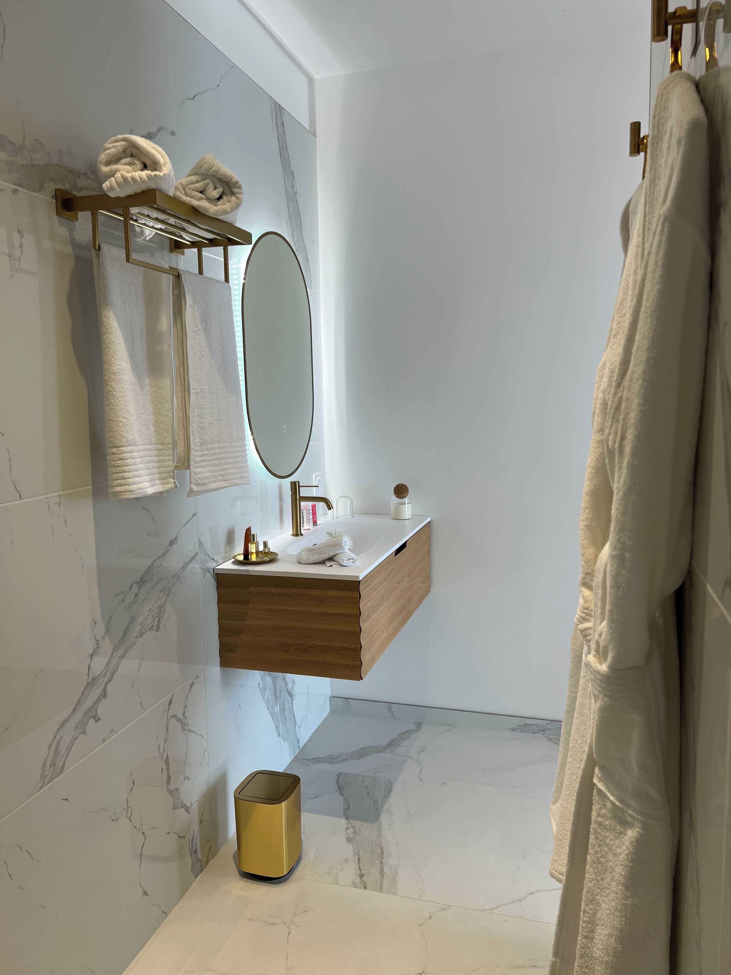 Un baño moderno con salpicadero de mármol, tocador de madera, cubo de basura dorado y un perchero con dos toallas.