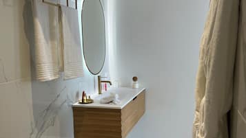 Un baño moderno con salpicadero de mármol, tocador de madera, cubo de basura dorado y un perchero con dos toallas.