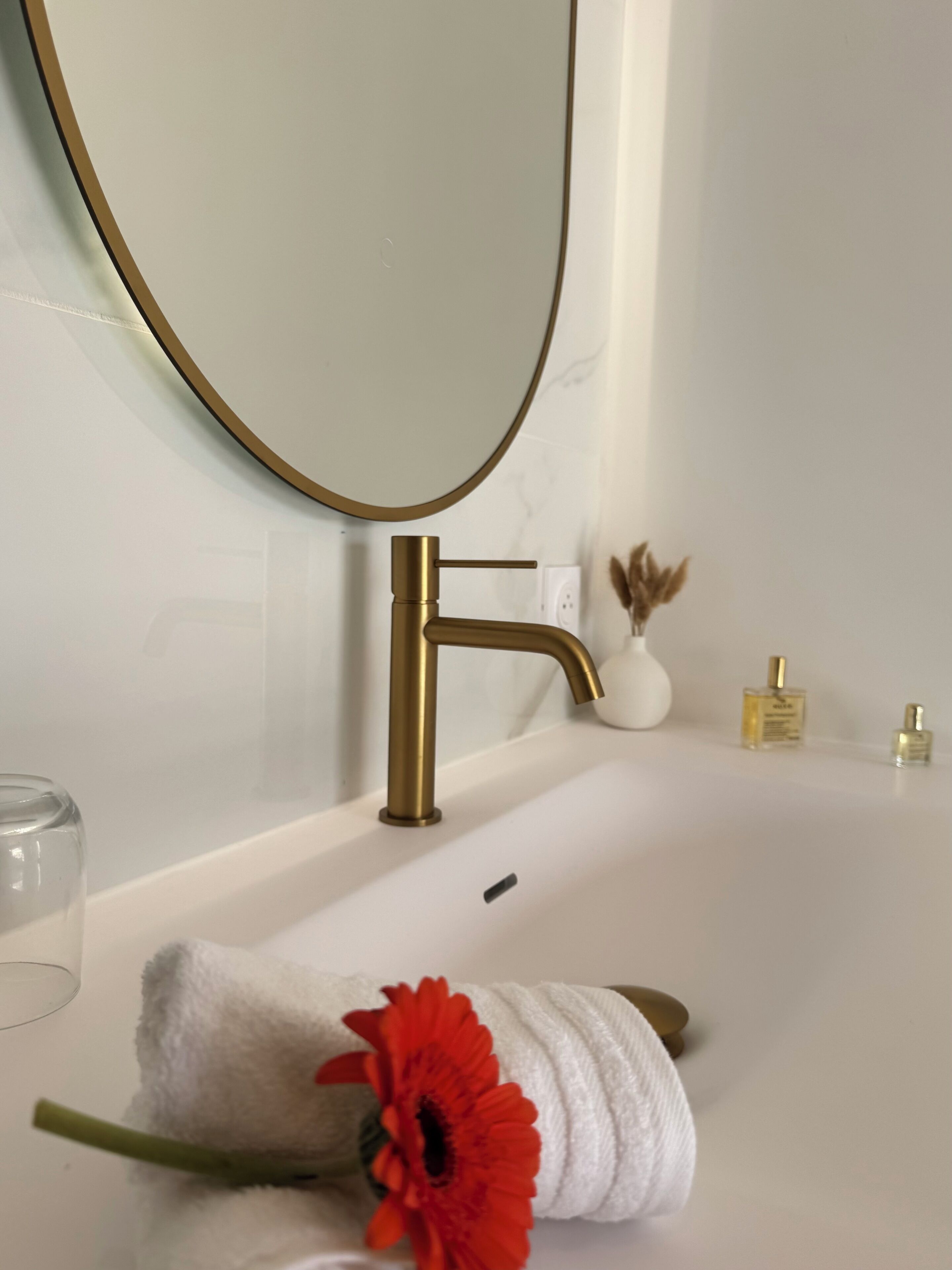 A bathroom with a white bathtub, a gold faucet, a round mirror, and a towel with a red flower.