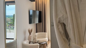 Une salle de bain moderne avec un peignoir blanc suspendu à un cintre doré, un miroir rond, un téléviseur à écran plat fixé au mur et une vue sur l’extérieur grâce à une grande fenêtre.
