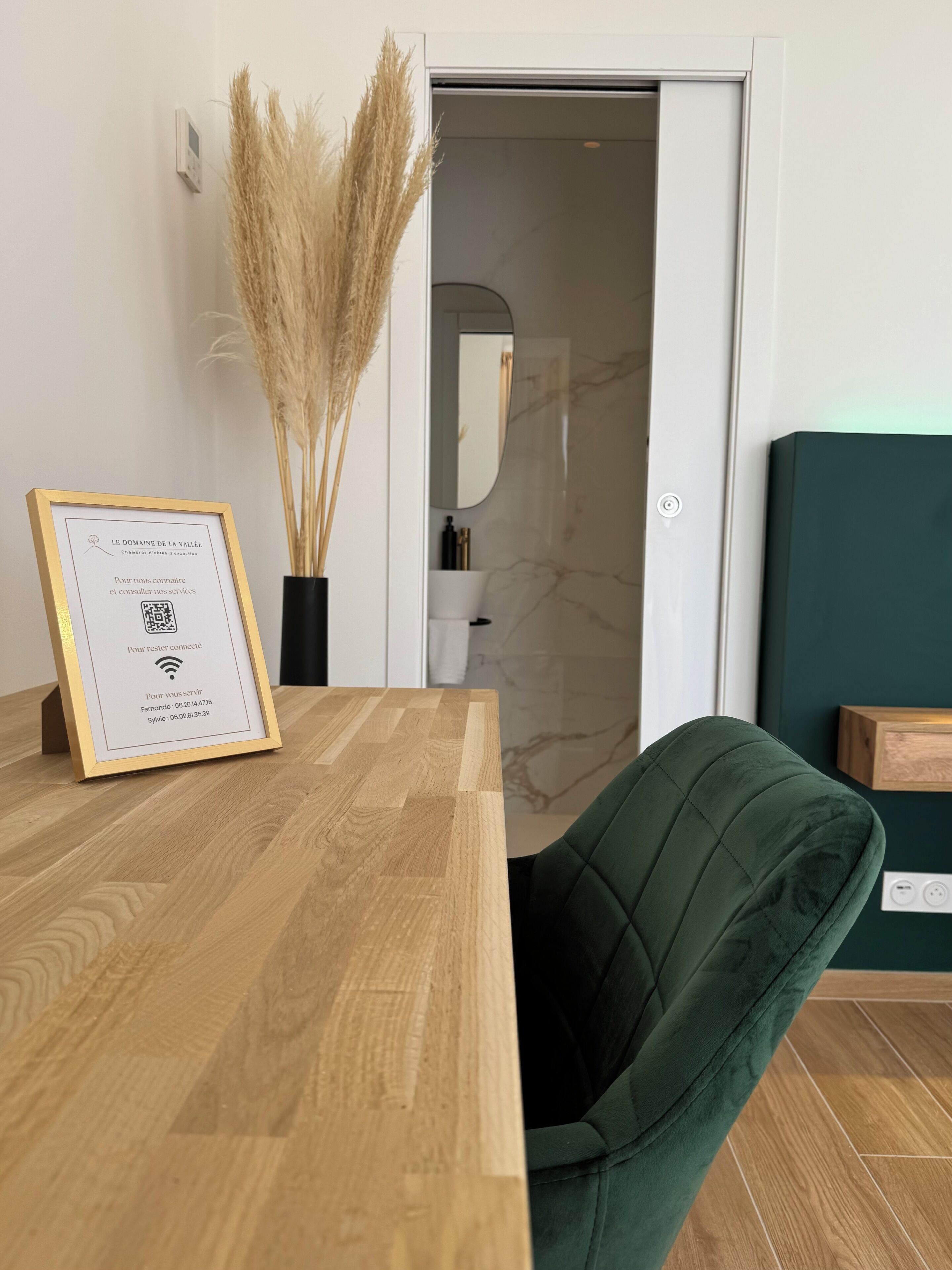 Un mostrador de recepción con un cartel enmarcado, un florero con plantas secas y una silla tapizada de verde.
