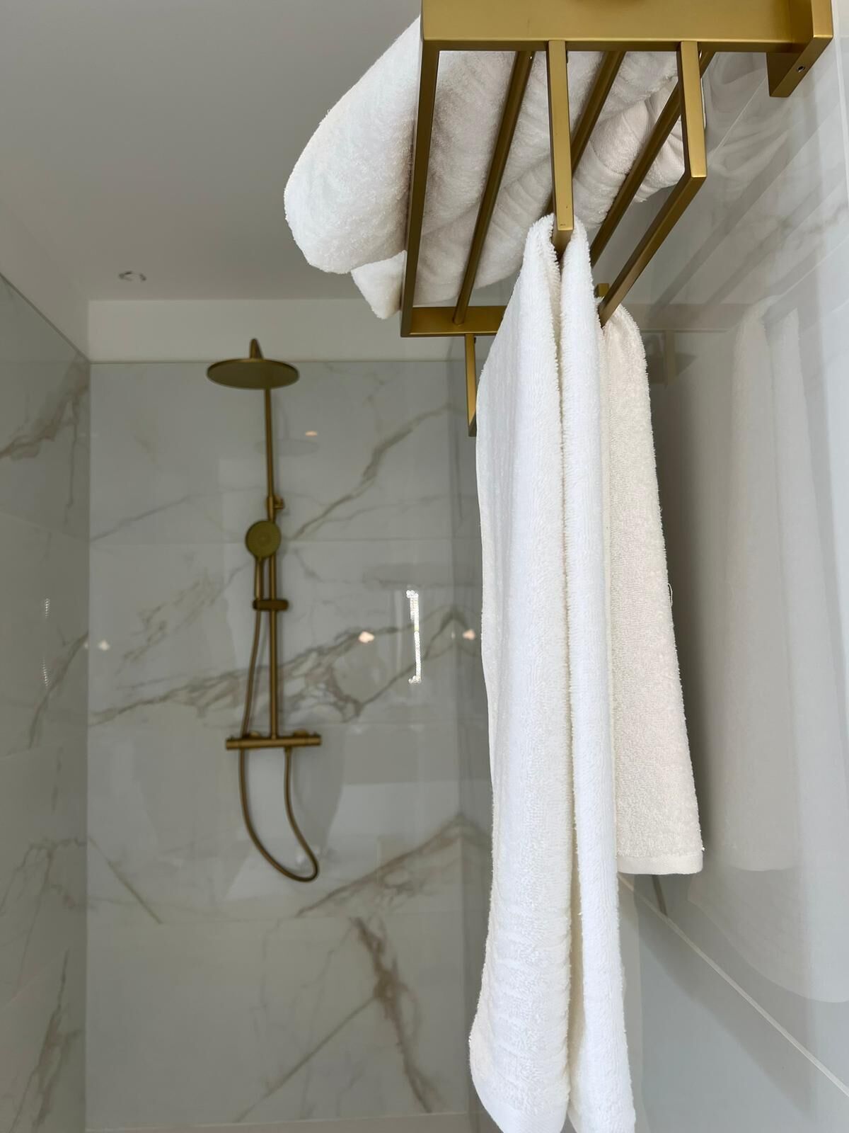 A modern bathroom with a marble shower wall, a gold showerhead, and white towels hanging on a gold towel rack.