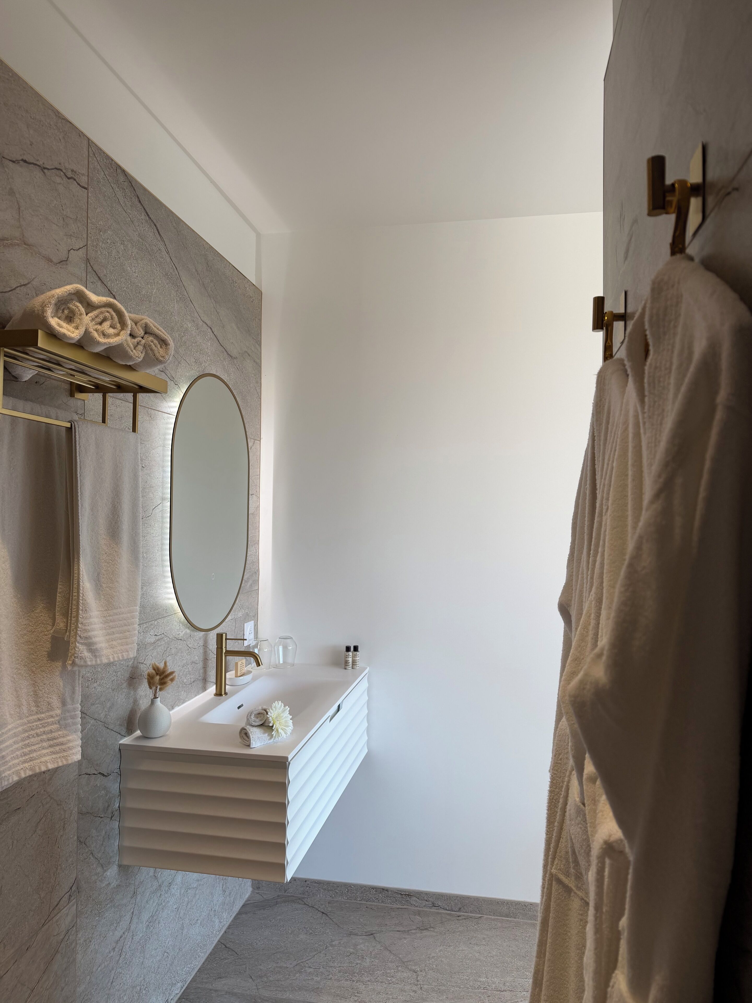 A modern bathroom with a marble wall, a gold-framed mirror, a white vanity, and a towel rack.