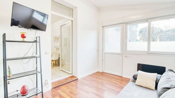 Una sala de estar moderna con un televisor de pantalla plana montado en la pared, un estante de vidrio con objetos decorativos, piso de madera y un ventanal que proporciona luz natural.