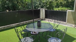 Un patio con una mesa redonda y sillas, rodeado de césped artificial verde, una cerca de metal y árboles al fondo.