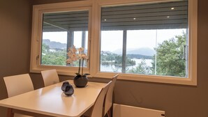A dining area with a table and chairs, a vase with flowers, and a view of a lake and mountains through large windows.