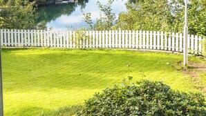 A view of a lake with a white picket fence in the foreground, surrounded by greenery and houses in the background.